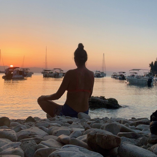 Anita sitting in a meditation pose on a rocky beach at sunset, looking out over calm water.
